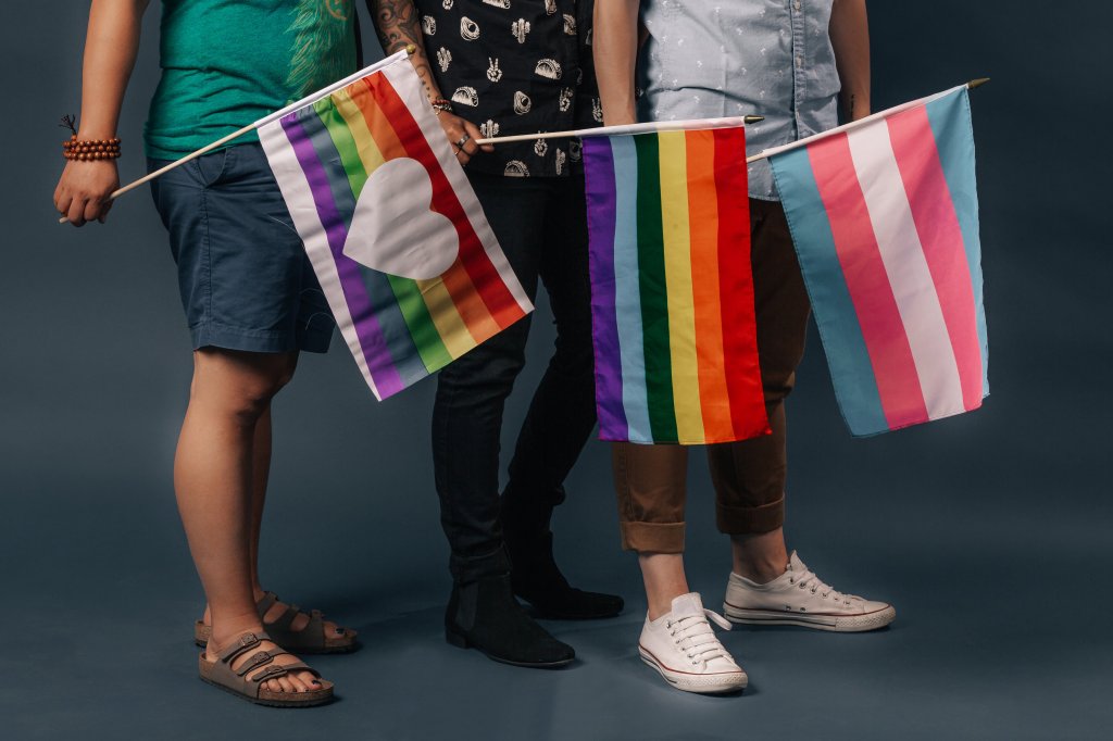 Three people standing, visible from the waist down. Each one holds a different Pride flag.