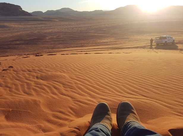 Person sitting on top of a sand dune, only their boots visible as they look across the desert to the setting sun.